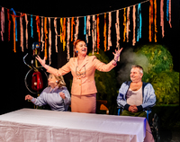 Zdjęcie ze spektaklu "Błoto". Przedstawia trzech aktorów przy stole zasłanym białym obrusem. Kobieta w środku stoi z rozłożonymi rękoma. Na ścianie za aktorami aplikacja z pociętej bibułki.