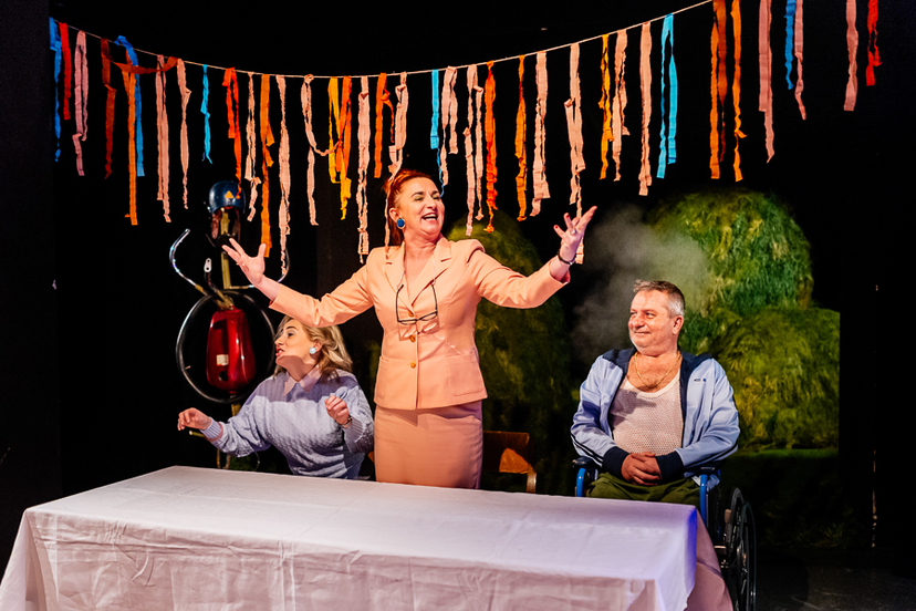 Zdjęcie ze spektaklu "Błoto". Przedstawia trzech aktorów przy stole zasłanym białym obrusem. Kobieta w środku stoi z rozłożonymi rękoma. Na ścianie za aktorami aplikacja z pociętej bibułki.