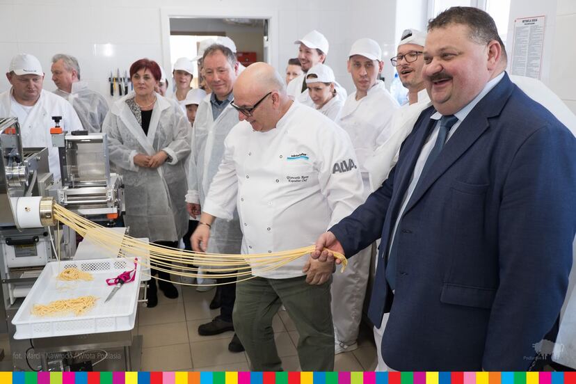 Wicemarszałek województwa wyciąga z maszyny pęk nitek makaronowych z pomocą włoskiego mistrza kuchni i w otoczeniu pracowników zakładu.