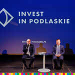 Marszałek Artur Kosicki i dyrektor Dąbrowski siedzą przy stole na tle ekranu. Na ekranie wyświetlony jest napis Invest in Podlaskie przy stoliku.  W tle niebieski ekran z napisem Invest in Podlaskie. 