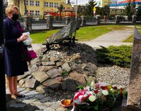 Kobieta trzyma znicz stojąc przed tablicą pamiątkową. Przed nią leżą kwiaty. W tle miejskie zabudowania.