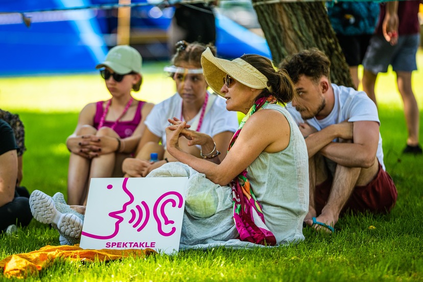 Grupa ludzi siedzi na trawie. Prze nimi stoi tabliczka z rysunkiem jak postać szepcze coś do ucha.