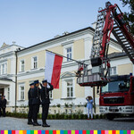 Trzech strażaków salutuje przed sztandarem biało-czerwonym zawieszonym na dźwigu