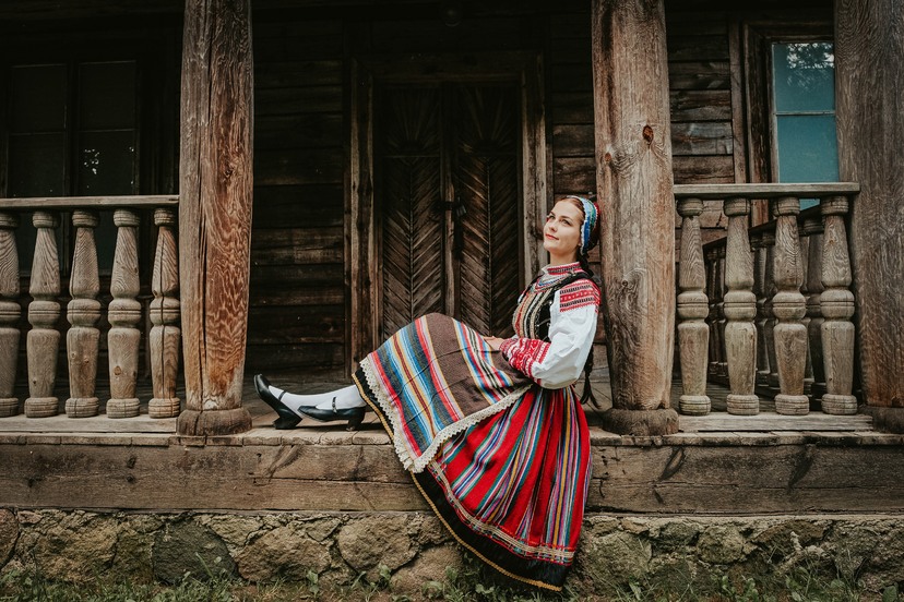 Dziewczyna ubrana w strój ludowy siedząca na ganku drewnianego domu