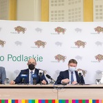 Trzech mężczyzn i jedna lobieta siedzą przy stole, na tle białego baneru z logo Województwa Podlaskiego