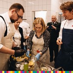 Radny Sebastian Łukaszewicz wraz z dwoma kobietami szykują jedzenie i pochylają się. Po prawej stronie stoi Karol Okrasa. W oddali komórkę trzyma mężczyzna w mundurze