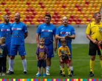 Kilku piłkarzy oraz sędzia ubrany na żółto stoją na boisku w jednym szeregu, Przed nimi stoją dzieci