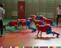 Dzieci w czerwonych koszulkach i niebieskich spodenkach ćwiczą na sali gimnastycznej