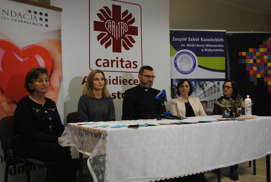Pięć osób siedzących za stołem na tle logo Caritas Archidiecezji Białotockiej