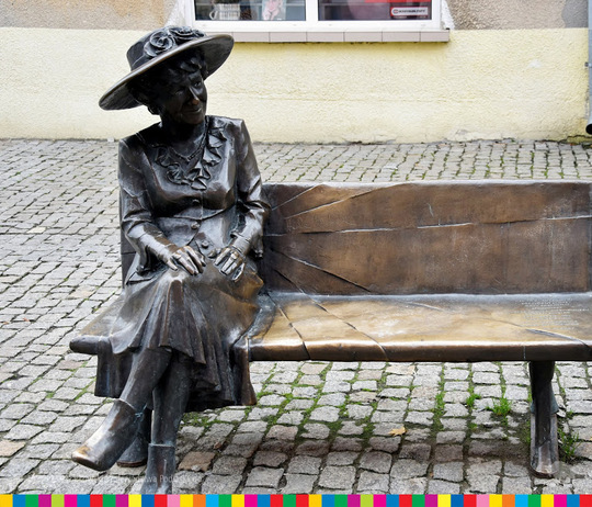 Metalowa ławeczka z siedzącą kobietą w kapeluszu.