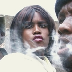 na zdjęciu czarnoskóra kobieta i dwóch mężczyzn, jeden z nich wypuszcza papierosowy dym z ust