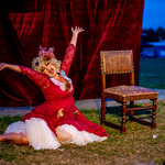 Aktorka przebrana w styl barokowy, w czerwonej sukni i peruce na głowie, siedzi na ziemi i przeciąga się.