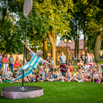 Spektakl w parku na trawie, wokół publiczność, pośrodku aktor w biało-błękitnym stroju cyrkowym podczas tańca przy rurze.