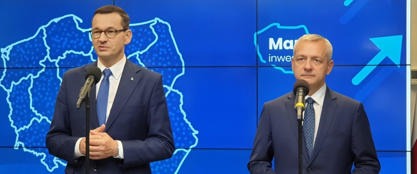 Premier M. Morawiecki i minister M. Zagórski stoją przed mikrofonami, w tle ekran z  białą grafiką mapy Polski na niebieskim tle.
