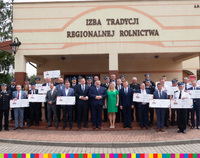 Samorządowcy i strażacy OSP w grupie na schodach przed wejściem do Izby Tradycji Regionalnej Rolnictwa w Nowych Piekutach - w rękach trzymają duże, symboliczne czeki z kwotami dotacji.