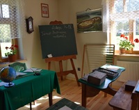 Sala z wystrojem dawnej klasy szkolnej: liczydło, stara tablica na sztalugach, stół z zielonym suknem a ana nim stare , szkolne dzienniki. Napis na tablicy: Lekcja, temat: Powtórzenie materiału