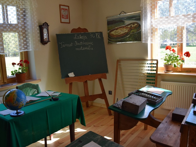 Sala z wystrojem dawnej klasy szkolnej: liczydło, stara tablica na sztalugach, stół z zielonym suknem a ana nim stare , szkolne dzienniki. Napis na tablicy: Lekcja, temat: Powtórzenie materiału