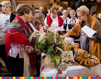 Kilka osób, w tym kobiety ubrane w stroje ludowe stoją przy stole z żywnością i wyrobami ludowymi.