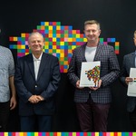 Czterech mężczyzn na ciemnym tle z kolorowymi pixelami. Dwóch z nich trzyma w ręku papierowe teczki
