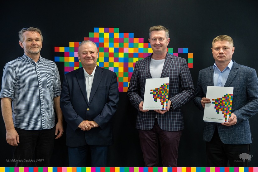 Czterech mężczyzn na ciemnym tle z kolorowymi pixelami. Dwóch z nich trzyma w ręku papierowe teczki