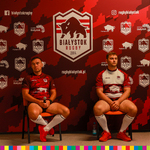 Dwóch zawodników siedzących pod ścianką z logo klubu oraz patronami