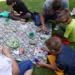 Dzieci siedzą przy kocu na, którym leżą kamienie. Jeden chłopiec maluje farbkami na białej kartce.