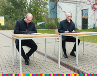 Marszałek Artur Kosicki oraz drugi mężczyzna siedzący przy stolikach na zewnątrz i podpisujący dokumenty