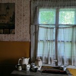 Na starym stoliku stoi stary dzbanek i naczynia, za nimi widok na stare okno, po lewej stronie na ścianie wisi obraz