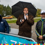 Trzech mężczyzn stoi za barierką reklamową. Stojący w środku trzymający parasol mówi do mikrofonu.