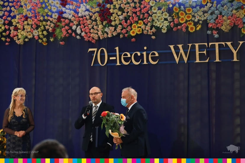 Marszałek Artur Kosicki mówiący do mikrofonu. Obok stoi wicemarszałek Marek Olbryś trzymający kwiaty. W tle napis 70-lecie WETY.