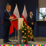 Członek zarządu województwa podlaskiego Marek Malinowski mówiacy do mikrofonu. Za nim stoi drugi mężczyzna oraz flagi Polski.