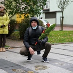 Mężczyzna kuca nad chodnikiem trzymając bukiet kwiatów. W tle stojące dwie osoby