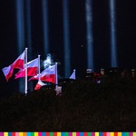 Flagi biało-czerwone na wzgórzu. W tle widoczne wiązki światła. Widoczne oddziały umundurowane Wojska Polskiego.