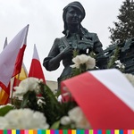 Widoczny pomnik kobiety w mundurze przy którym widnieją flagi biało-czerwone oraz kwiaty.