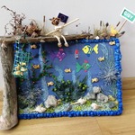 Praca, która przedstawia ryby, kukiełkę z wędką, rośliny, kamienie. Tło pracy niebieskie.