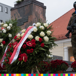 Wieńce z białoczerownych kwiatów leżące pod pomnikiem i obok stojącego żołnierza