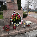 Wieniec z z kwiatów, dwa kwiatki w doniczkach oraz dwa znicze stojące pod pomnikiem. Wszystko w biało czerwonych barwach