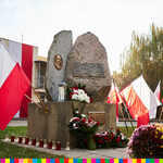 Tablica pamiątkowa umieszczona na pomniku w kształcie kamienia. Obok znajdują się kwiaty, znicze oraz flagi biało-czerwone. 