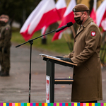Mężczyzna mówiący do mikrofonu. Na płaszczu ma godło Polski. Za nim stoją flagi Polski.