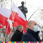 Na pierwszym planie widocznych dwóch mężczyzn na tle biało-czerwonych flag oraz pomnika.