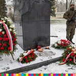 Obok cokołu pomnika stoi wieniec z biało-czerwoną wstęgą. Widoczne są zapalone znicze oraz kwiaty związane biało-czerwonymi wstążkami. Z prawej strony stojący na baczność żołnierz trzymający karabin