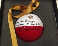 Biało-czerwona bombka z godłem Polski z zieloną wstążką leżąca w pudełku, na której widnieje napis "Dla dzielnego Rycerza Mikołaja M. Morawiecki"