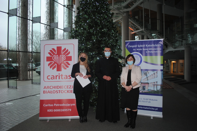 Trzy osoby stoją na tle choinki. Po lewej baner Caritasu, po prawej - Zespołu Szkół Katolickich.