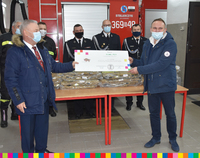 Wicemarszałek Marek Olbryś wręcza mężczyźnie symboliczny czek. Na drugim planie stoi kilku strażaków ubranych w mundury na tle wozu strażackiego.