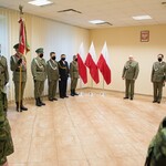 Grupa umundurowanych funkcjonariuszy Straży Granicznej stoi wokół pocztu sztandarowego. Obok trzy flagi biało - czerwone i makieta słupka granicznego w kolorze biało - czerwonym z godłem Polski.
