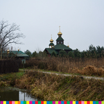 Wśród łąk, za zbiornikiem wodnym i ogrodzeniem widoczna cerkiew z dwiema wieżami ze złotymi kopułami.