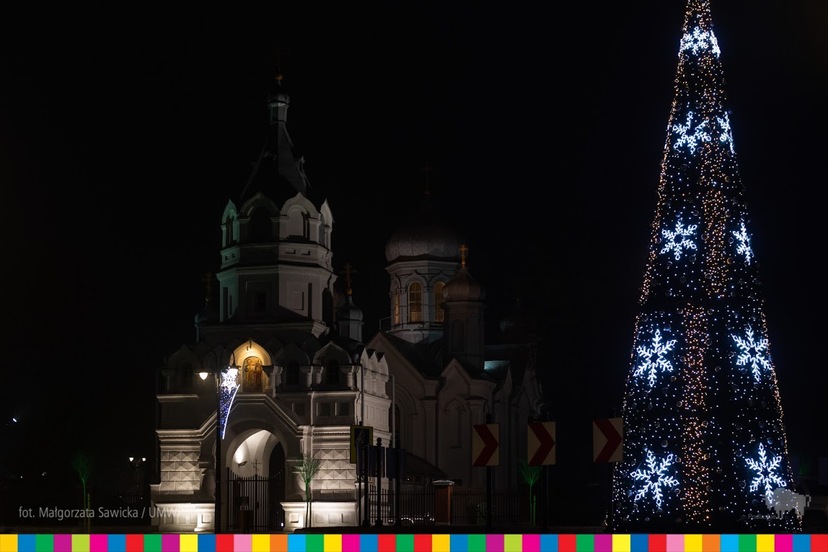 Po prawej choinka oświetlona lampkami w kształcie gwiazdek. Po lewej podświetlony kościół.
