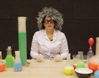 Kobieta w szarej peruce na głowie oraz białym fartuchu siedzi przed rozstawionymi naczyniami laboratoryjnymi