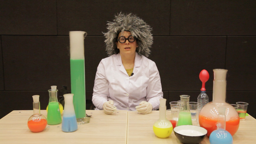 Kobieta w szarej peruce na głowie oraz białym fartuchu siedzi przed rozstawionymi naczyniami laboratoryjnymi