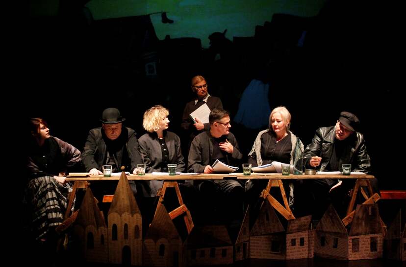 Aktorzy siedzą za dużym stołem. Za nimi scenografia teatralna przedstawiająca miasteczko.